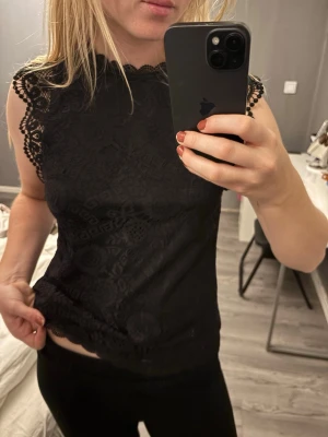 Svart spetsblus med mönster - Säljer en svart blus med vackra spetsdetaljer och diskret mönster. Toppen är ärmlös med rundad hals och har fina spetskanter vid axlarna.