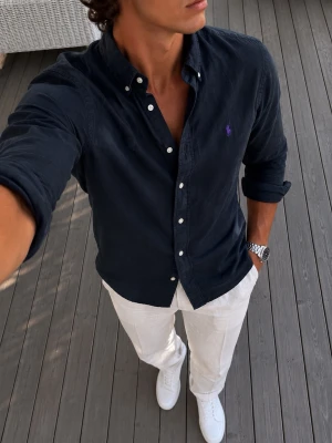 Svart skjorta från Ralph Lauren - Klassisk svart skjorta från Ralph Lauren med button-down krage och vita knappar. Skjortan har lång ärm och är tillverkad i mjuk bomull. Liten broderad logga i lila på bröstet ger en stilren touch.