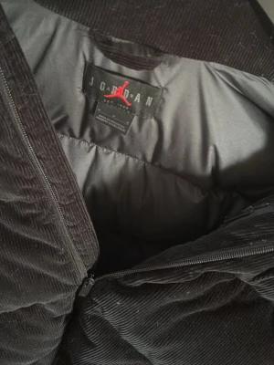 Svart pufferjacka från Jordan - Säljer en svart pufferjacka från Jordan med breda paneler och dragkedja framtill. Jackan är tillverkad i ett mjukt manchestertyg som ger en schysst textur och extra värme. Perfekt för kalla vinterdagar och har en klassisk, sportig look.