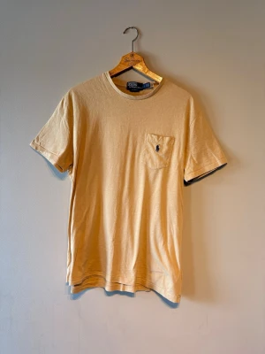 Beige t-shirt från Polo Ralph Lauren - Säljer en beige t-shirt från Polo Ralph Lauren med klassisk passform. T-shirten har korta ärmar, rund halsringning och en bröstficka med den ikoniska broderade loggan. Tillverkad i mjuk bomull för skön känsla.