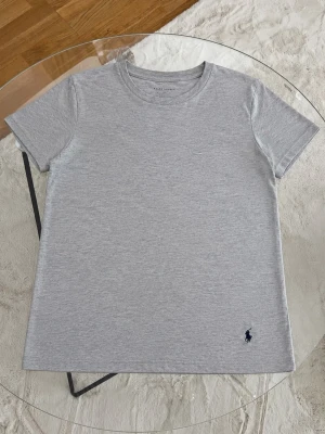 Grå t-shirt från Ralph Lauren - Snygg och enkel grå t-shirt från Ralph Lauren med klassisk passform och korta ärmar. Tillverkad i mjuk bomull och har den ikoniska lilla mörkblå loggan broderad nertill. Perfekt basplagg till garderoben.