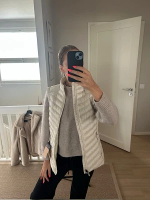 Ljusbeige dunväst med hög krage - Snygg ljusbeige dunväst med quiltad design och hög krage. Västen har dragkedja framtill och två sidofickor med dragkedja. Perfekt att slänga över en tröja för extra värme. Stilren och enkel att matcha till olika outfits.