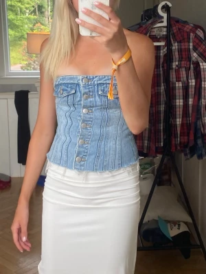 Ljusblå jeans topp med knappar - Trendig tubtopp i ljusblå denim med knappar framtill och dekorativa sömmar. Toppen är axelbandslös och har en croppad passform som ger en cool och modern look. Perfekt att matcha med kjol eller byxor för en snygg outfit.