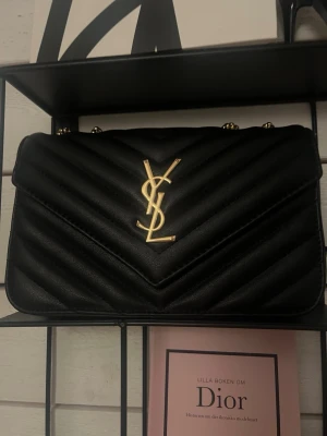 Svart YSL axelväska  - Säljer en svart axelväska från YSL i quiltat läder med guldfärgade detaljer och kedjerem. Väskan har det klassiska YSL-emblemet framtill och en elegant, tidlös design som passar till många olika looks.
