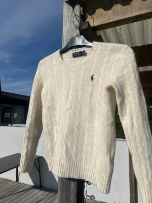 Vit kabelstickad tröja Polo Ralph Lauren - Kabelstickad vit tröja från Polo Ralph Lauren med rund halsringning och den klassiska broderade loggan på bröstet. Tröjan har långa ärmar och ribbade muddar vid ärmslut och nederkant. Perfekt för lager på lager under kyliga dagar.