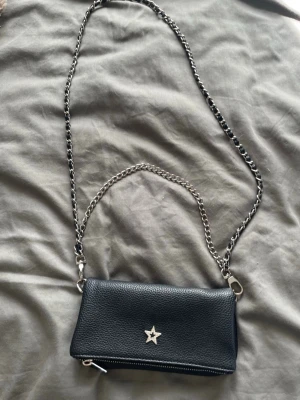 Svart axelväska med kedjerem och stjärna - Snygg svart axelväska i läderimitation med silverfärgad kedjerem och dekorativ stjärna framtill. Väskan har dragkedja upptill och magnetknappar på locket. Perfekt för att bära det viktigaste!