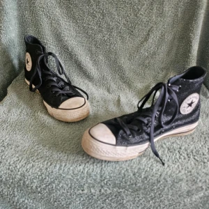 Svarta Converse Chuck Taylor Highs - Klassiska svarta Converse Chuck Taylor All Star high-top sneakers med vit tåhätta och sula. Skorna har svart tyg, vita detaljer och den ikoniska loggan på sidan. Perfekta för en avslappnad och cool stil.