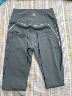Grå leggings från Alo med fickor - Snygga grå leggings från Alo med hög midja och praktiska sidofickor. De har vita kontrastsömmar och är tillverkade i ett stretchigt, mjukt material som passar perfekt till träning eller chill. Tight passform och modern look.