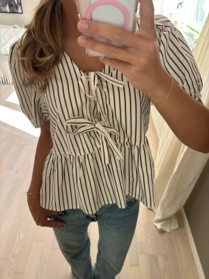 Randig blus med knyt och volang - Säljer en vit blus med svarta vertikala ränder och två dekorativa knyt framtill. Blusen har korta puffärmar och volang nedtill för en söt och trendig look. Perfekt till jeans eller kjol för en avslappnad men stilsäker outfit.