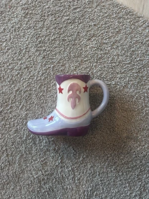 Mugg formad som cowboyboot - Unik mugg i keramik formad som en cowboyboot i lila, vitt och rosa med röda stjärnor. Handtaget sitter på skaftet och muggen har detaljer som påminner om westernstil. Perfekt för dig som gillar annorlunda muggar eller vill ha något kul till köket.