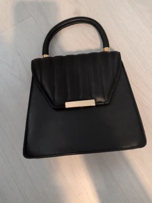 Svart handväska med gulddetaljer - Snygg svart handväska i konstläder med guldfärgade detaljer och kedjerem. Väskan har ett quiltat lock och stilren, fyrkantig form. Perfekt för dig som gillar klassisk och trendig design. Bärs i handtaget eller över axeln med kedjan.