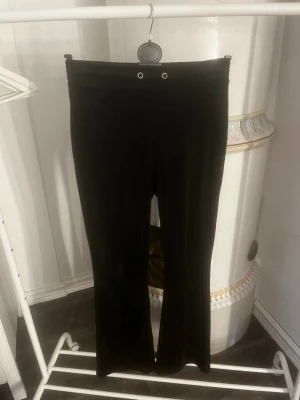 Svarta mjukis från Gina - Säljer ett par svarta mjukis med utsvängda ben och hög midja. De har två silverfärgade öljetter framtill i midjan för en cool detalj. Materialet ser stretchigt och mjukt ut, perfekt för en avslappnad och trendig look.