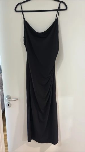 Svart långklänning med tunna band - Stilren svart långklänning med tunna axelband och draperad detalj framtill. Klänningen har en mjuk, följsam passform och ett elegant fall. Perfekt för dig som gillar minimalistisk och tidlös stil.
