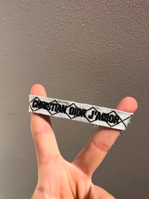 Grått tygarmband med svart text - Säljer ett grått tygarmband med svart broderad text och grafiskt mönster. Armbandet har en bred design och avslutas med fransar. Perfekt för dig som gillar streetwear och unika accessoarer.