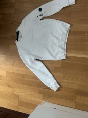 Vit sweatshirt från C.P. Company - Vit sweatshirt från C.P. Company med rund hals och ribbade muddar. Tröjan har en ikonisk svart detalj på ärmen med logotyp. Perfekt för en clean och stilren look.