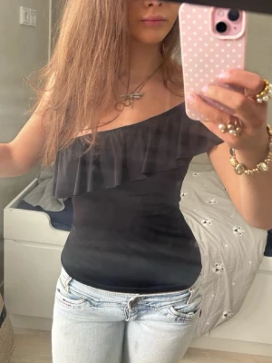Svart oneshoulder topp med volang - Snygg svart oneshoulder topp med volangdetalj längs ringningen. Toppen är tight och har en stilren look som passar perfekt till jeans eller kjol. Materialet känns mjukt och följsamt mot kroppen.