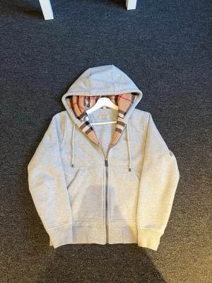 Grå Burrbery zip hoodie  - Snygg grå hoodie med dragkedja och klassisk huva. Insidan av huvan har ett rutigt mönster i beige, svart och rött som ger en unik touch. Hoodien har fickor framtill och ribbade muddar vid ärmar och nederkant. Perfekt för en chill och avslappnad stil.