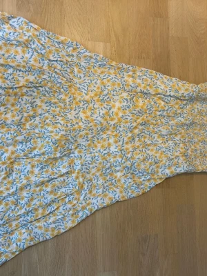 Blommig långklänning - Söt långklänning med blommigt mönster i gult och blått. Klänningen är luftig och har ett innertyg i vitt. Perfekt för varma dagar och har en romantisk känsla med sitt blommiga tryck och lätta material.