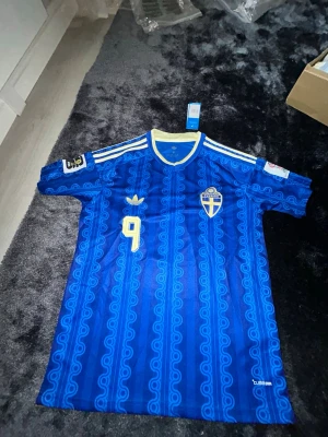 Alexander Isak nummer 9 storlek M - Säljer en blå fotbollströja från Sverige med nummer 9 på bröstet. Tröjan har gula detaljer, mönstrad design och Sveriges landslagsemblem på vänster sida. Klassiska Adidas-ränder på axlarna och korta ärmar. Perfekt för match eller träning.
