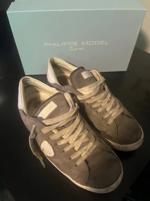 Grå Philippe Model sneakers - Snygga grå sneakers från Philippe Model med vita detaljer och logga på sidan. Skorna har en klassisk låg siluett, vita skosnören och är tillverkade i mocka och läder. Perfekta för dig som gillar stilrena och trendiga sneakers. Skorna är såklart äkta och Box följer med. 