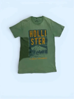 Grön Hollister t-shirt med tryck - Snygg grön t-shirt från Hollister med stort tryck framtill i gult och mörkblått. Motivet visar en palm och texten 'LA JOLLA CALIFORNIA'. Klassisk passform och rund halsringning. Perfekt för en avslappnad stil.
