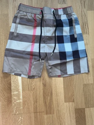 Beige badshorts från burberry - Snygga shorts med rutigt mönster i beige, svart, vitt och rött. Shortsen har elastisk midja med svart snörning och fickor på sidorna samt en bakficka. Perfekta för sommaren och har en avslappnad passform.(size m passar s)