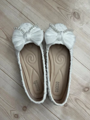 Vita ballerinaskor med rosett och pärlor - Säljer ett par vita ballerinaskor från CUCCOO som bara används en gång. Mycket bra skick