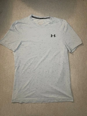 Ljusgrå Under Armour t-shirt - Snygg ljusgrå t-shirt från Under Armour med diskret mönster och logga på bröstet. Tillverkad i ett mjukt och lätt material som passar perfekt för träning. Klassisk passform och rund halsringning.