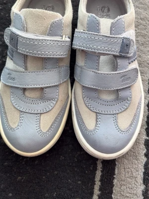 Grå sneakers med kardborre från Tino - Snygga sneakers i grått och beige mocka och skinn från Tino. Skorna har dubbla kardborreband för enkel påtagning och en vit sula. Baksidan har en blå detalj i mocka som ger extra stilpoäng. Perfekta för dig som gillar en clean och sportig look.
