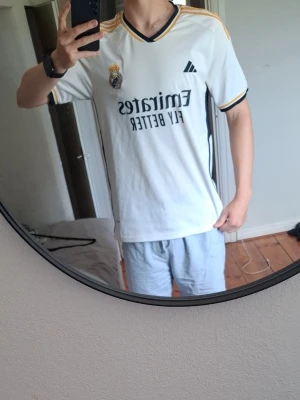 Real Madrid matchtröja Bellingham 5 - Säljer en vit Real Madrid matchtröja från Adidas med svarta och guldiga detaljer. Tröjan har klubbmärke och sponsortryck på bröstet samt BELLINGHAM och nummer 5 på ryggen. Perfekt för fotbollsfans som vill stötta sitt lag med stil.