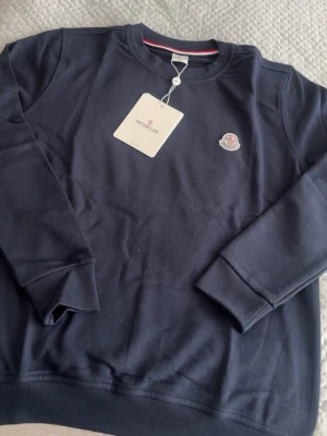 Mörkblå sweatshirt från Moncler - Snygg mörkblå sweatshirt från Moncler med rund halsringning och Moncler-logga broderad på bröstet. Tröjan har långa ärmar, ribbade muddar och är gjord i mjukt bomullsmaterial. Perfekt för en clean och stilren look.