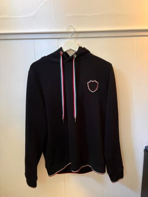 Svart italia hoodie 19V69 - Svart italia hoodie med huva och 🇮🇹detaljer. Har en liten sköldformad patch på bröstet och matchande färgdetaljer vid fållen. Perfekt för dig som vill ha en stilren hoodie med en snygg touch  