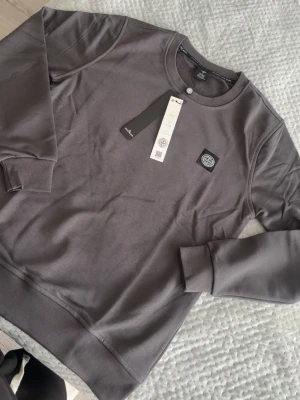 Grå sweatshirt från Stone Island - Snygg grå sweatshirt från Stone Island med rund halsringning och ribbade muddar. Tröjan har en diskret logopatch på bröstet och är tillverkad i mjuk bomull för maximal komfort. Perfekt för en clean och stilren look.