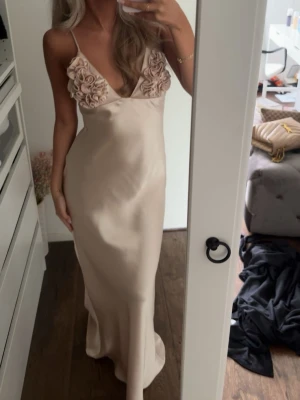 Champagnefärgad långklänning i satin - Elegant långklänning i champagnefärgad satin med smala axelband och djup v-ringning. Klänningen har dekorativa blommor vid bysten och en figurnära passform som ger en lyxig känsla.