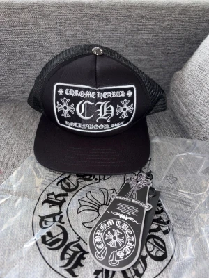 Chrome Hearts truckerkeps svart - Svart truckerkeps från Chrome Hearts med vit broderad logga och detaljer framtill. Kepsen har mesh på sidorna och justerbar snapback baktill. Klassisk streetwear-stil med tydliga Chrome Hearts-motiv och etiketter.