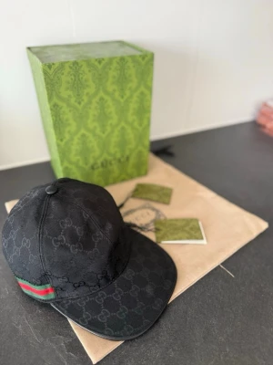 Gucci keps  - Svart keps från Gucci med klassiskt GG-monogram över hela kepsen. På sidan finns en ikonisk grön och röd rand. Kepsen har böjd skärm och justerbar rem baktill. Materialet är textil och kepsen levereras med originalförpackning och tags.