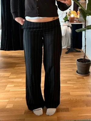 Svarta kostymbyxor med vita ränder - Säljer ett par svarta kostymbyxor med vita tunna kritstreck. Byxorna har en rak och lite vidare passform med låg midja. Klassisk look som funkar till många olika stilar. Materialet ser ut att vara en blandning med polyester för en mjuk känsla.