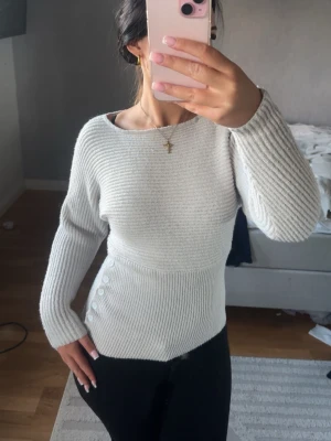 Vit ribbstickad tröja med knappar - Snygg vit ribbstickad tröja med lång ärm och rund halsringning. Tröjan har en figurnära passform med dekorativa knappar på sidan av midjan som ger en unik touch.