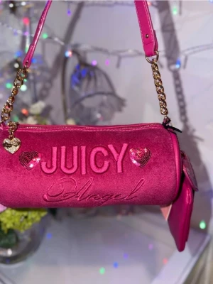 Rosa Juicy Couture axelremsväska - Trendig rosa axelremsväska från Juicy Couture med broderad text 'JUICY Angel' och paljettdekorerade hjärtan. Väskan har guldfärgade kedjedetaljer, hjärtberlock och matchande liten accessoarväska. Materialet är mjuk velour som ger en lyxig känsla.