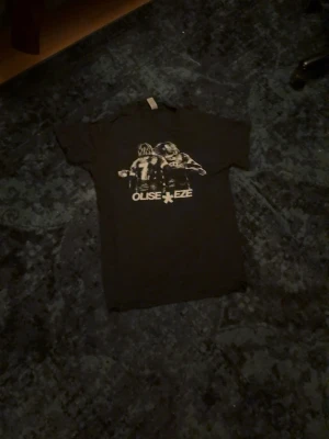 Svart t-shirt med Olise & Eze tryck - Cool svart t-shirt med ett stort tryck på ryggen som visar två fotbollsspelare och texten 'OLISE EZE'. T-shirten har klassisk passform. Perfekt för dig som gillar sportiga och unika plagg.