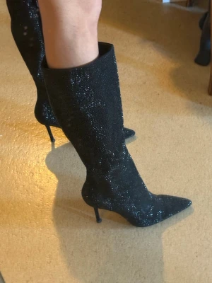 Svarta glittriga stövletter med klack - Säljer ett par svarta stövletter med smal hög klack och spetsig tå. Skorna är täckta av glittrande detaljer som ger en riktigt snygg look. De går upp till halva vaden och har dragkedja på insidan. Nubben på klacken är lite sliten men går att byta ut! 7cm klack i höjd!