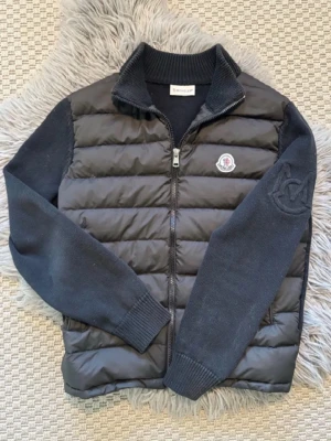 Moncler Cardigan - Snygg svart dunväst från Moncler med stickade ärmar och ribbad krage. Västen har quiltad front, dragkedja och Moncler-logga på bröstet. Perfekt för lager-på-lager och ger en clean look med exklusiv känsla.