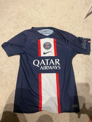 PSG matchtröja blå Nike - Säljer en blå PSG matchtröja från Nike med vita och röda detaljer. Tröjan har klubbmärke och Qatar Airways-tryck på bröstet samt korta ärmar. Perfekt för fotbollsträning eller att bära till vardags. Materialet är lätt och andas bra.