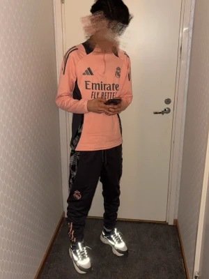 Real Madrid träningsset rosa/svart - Säljer ett snyggt Real Madrid träningsset från Adidas. Tröjan är långärmad i rosa med svarta detaljer och klubbmärke på bröstet. Byxorna är svarta med rosa detaljer och klubbmärke på benet. Perfekt för fotbollsträning eller chill.
