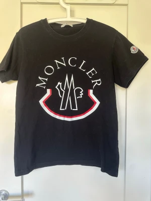 Svart Moncler t-shirt med stor logga - Snygg svart t-shirt från Moncler med stor vit och röd logga framtill och Moncler-märke på ärmen. Klassisk rund hals och korta ärmar. Perfekt för dig som vill ha en stilren och trendig look.