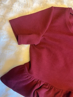 Vinröd topp med volangkant - Snygg vinröd topp med rund halsringning och korta ärmar. Toppen har en volangkant nedtill som ger en extra touch till looken. Materialet känns mjukt och har en lätt struktur. Perfekt att styla med jeans eller kjol för en trendig outfit. Tröjan säljer jag därför att jag tycker den är för kort vid magen/ryggen. Jag är ca 160cm och den slutar strax under nacken för mig. Tröjan är i xs och skulle garanterat passa någon som vanligtvis är en xs!