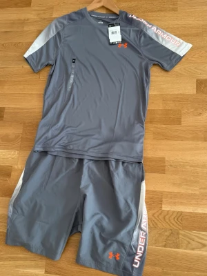 Grått träningsset från Under Armour - Helt nytt med prislapp på. Tröjan är i storlek S, shortsen storlek L men passar även M. Priser kan diskuteras vid snabbt köp. Snyggt träningsset från Under Armour i grått med vita detaljer och orange logga. Setet består av en t-shirt och shorts i snabbtorkande funktionsmaterial, perfekt för gymmet eller löpning. Under Armour-tryck på ärm och ben ger en sportig look.