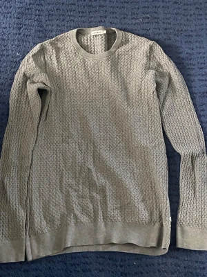 Grå stickad tröja från J.Lindeberg - Snygg grå stickad tröja från J.Lindeberg i storlek M. Tröjan har ett diskret strukturmönster, rund halsringning och ribbade muddar vid ärmslut och nederkant. Perfekt till höst och vinter för en stilren look.
