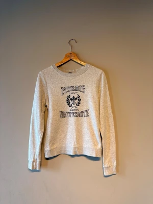 Grå sweatshirt Morris Lady Université - Snygg ljusgrå sweatshirt från Morris Lady med trycket 'Université Paris' och en svart dekorativ lagerkrans på bröstet. Tröjan har rund halsringning, långa ärmar och ribbade muddar. Perfekt för en avslappnad och trendig look.