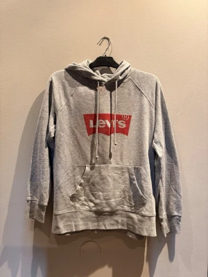Grå hoodie från Levi's med logga - Säljer en grå hoodie från Levi's med den klassiska röda loggan på bröstet. Hoodien har huva med dragsko och en stor magficka. Tillverkad i mjukt sweatshirtmaterial och har ribbade muddar vid ärmar och nederkant. Perfekt för en chill och avslappnad stil.